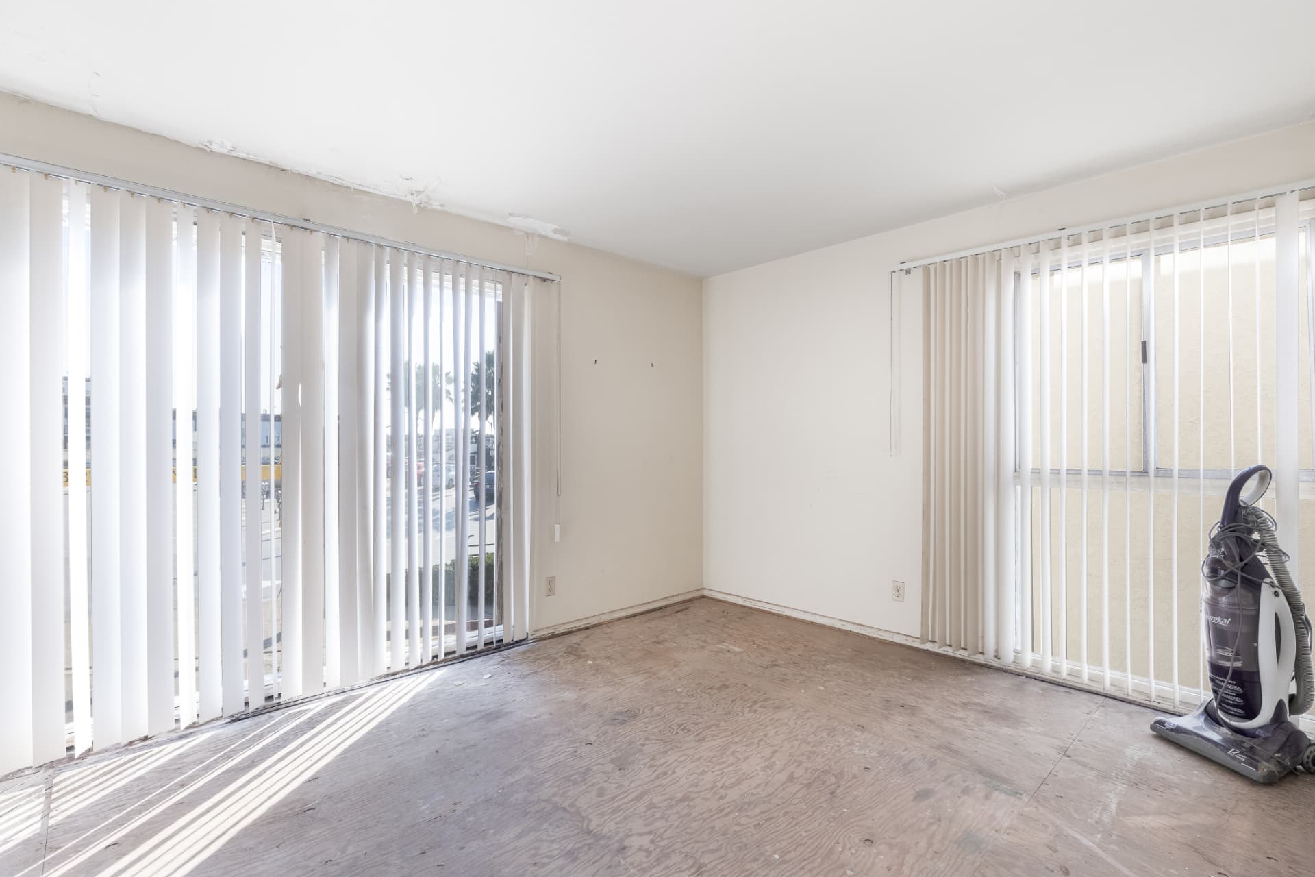 Before: Living room before renovation with bare subfloor and vertical blinds